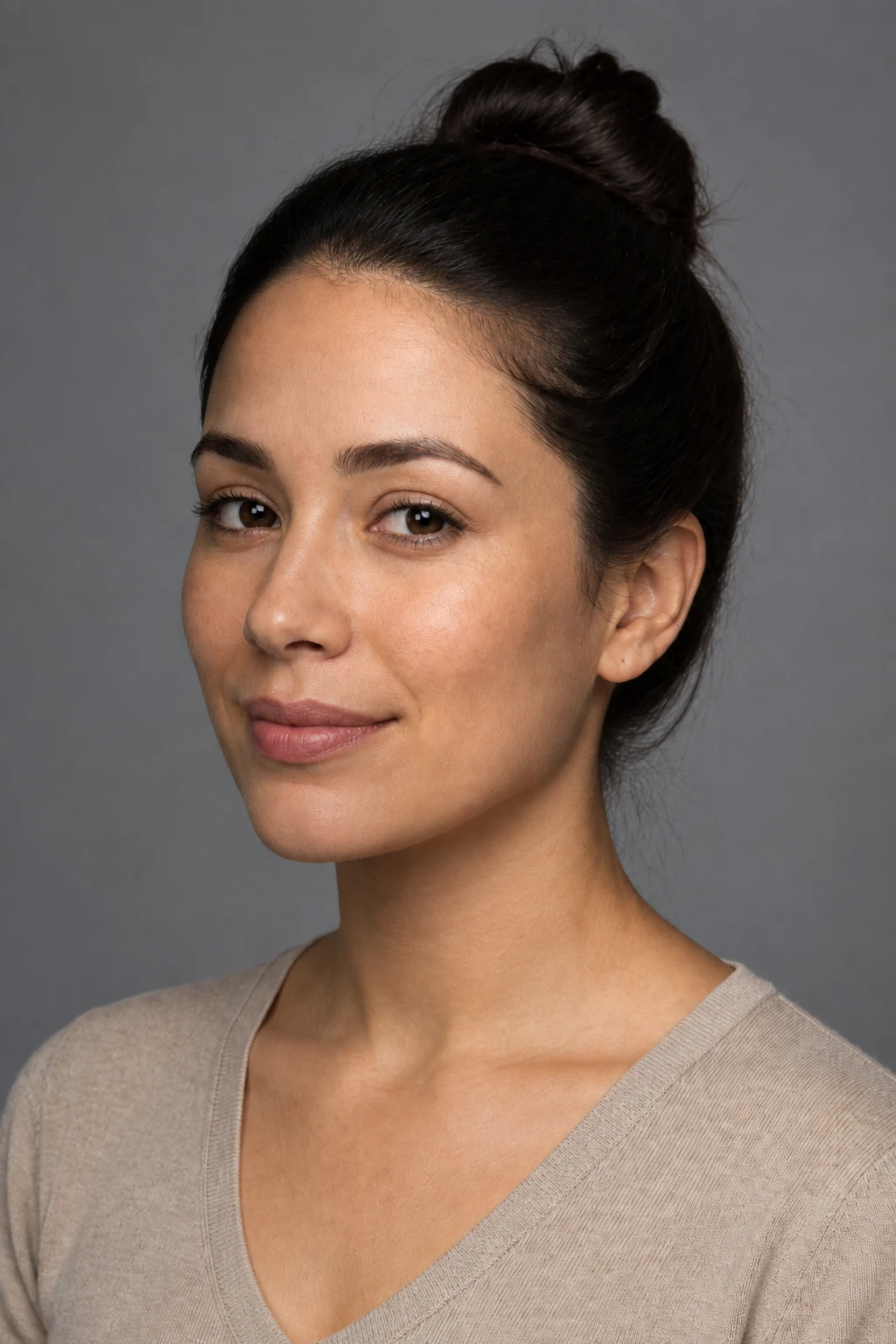 White fill lifts the near cheek about a stop while the softbox stays camera-left on the far cheek — softer short light… Woman with hair in a bun in a tan sweater on grey, softbox still camera-left on the far cheek while white fill opens the near cheek — softer short light, not broad.